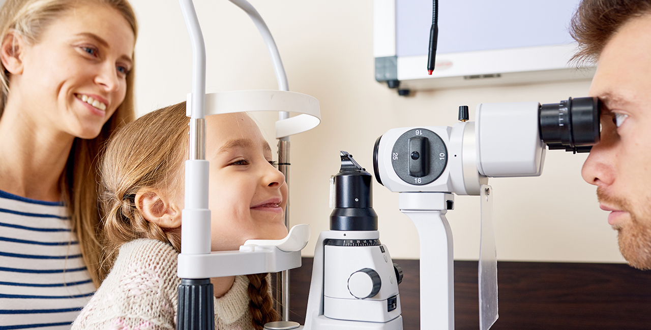 Portrait of cute little girl smiling happily looking at slit lamp machine during medical check up in eye clinic